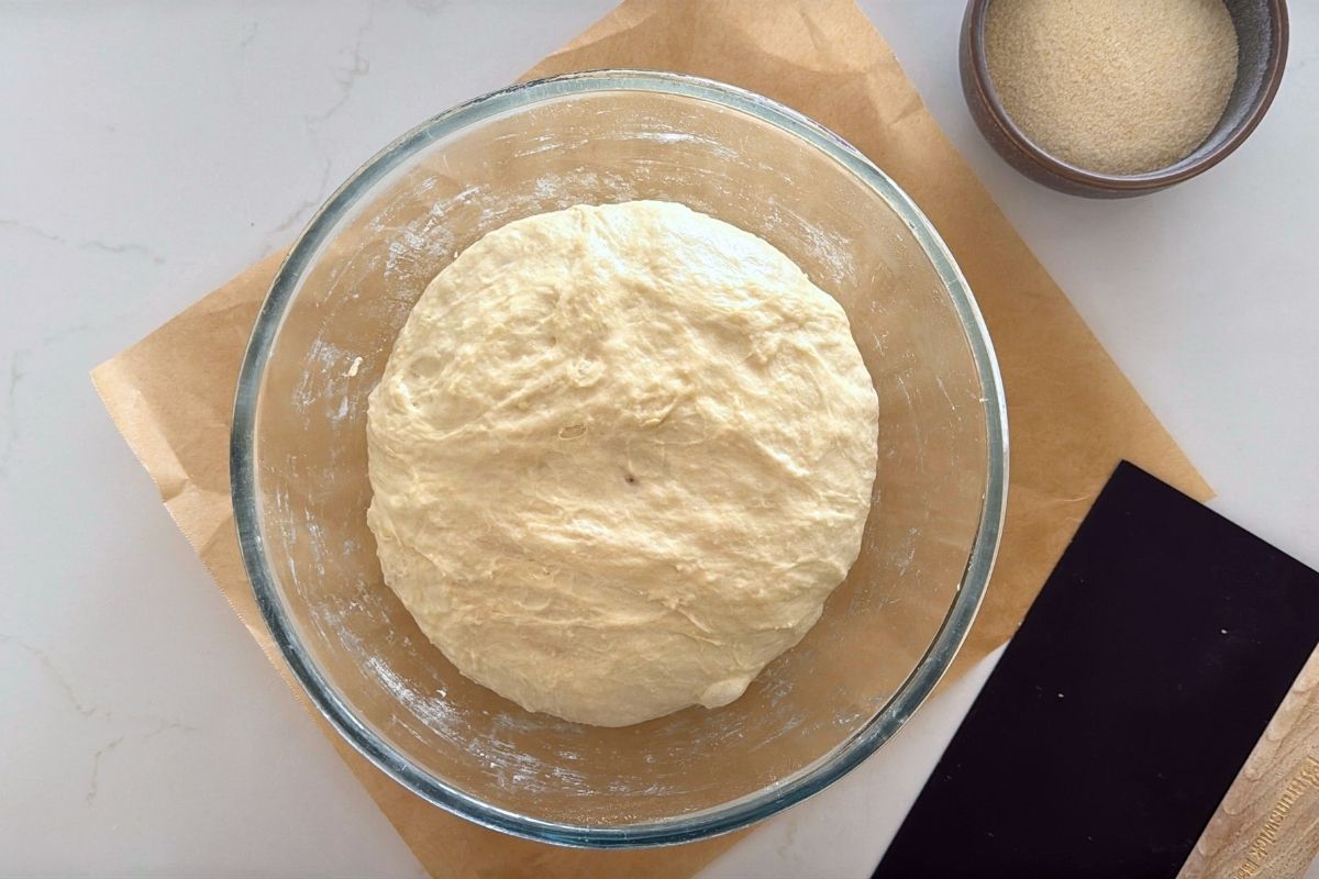 A glass bowl sitting on a square piece of parchment paper. Inside the bowl, the dough has doubled and is ready to shape.