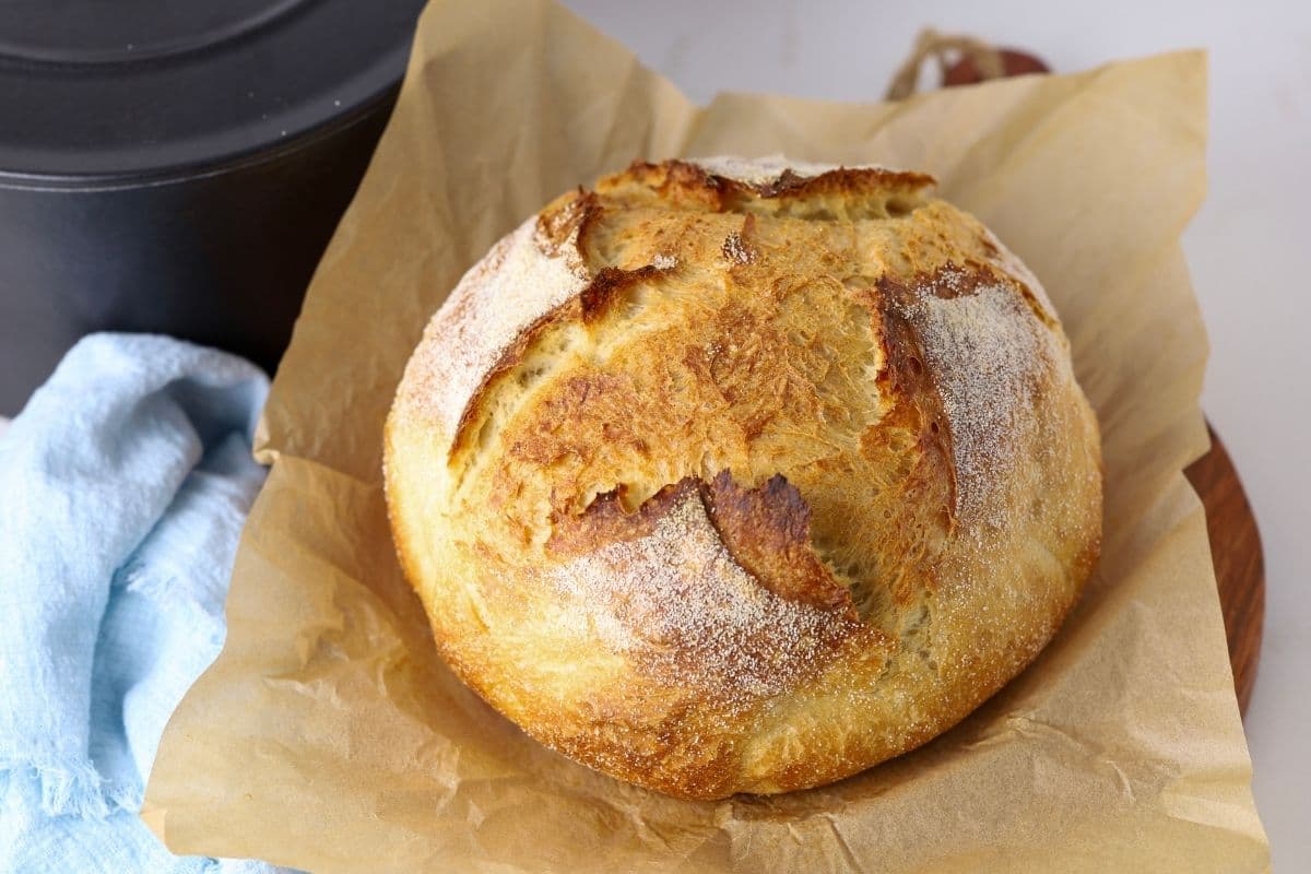 A crusty loaf of sourdough discard bread sitting on a piece of parchment paper. The loaf has been scored with a simple cross on top and you can see a pale blue linen napkin next to the loaf.