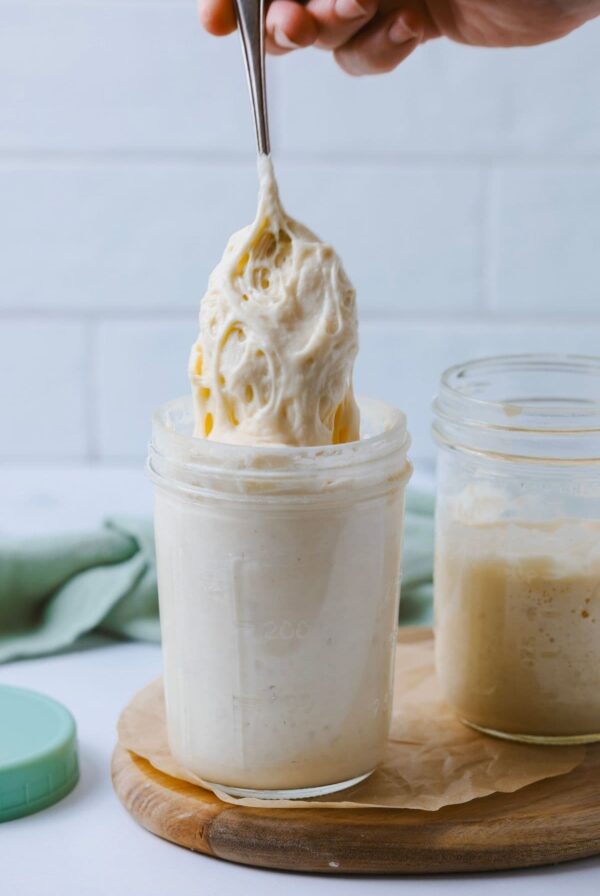A photo of a strong, bubbly sourdough starter being lifted out of a glass jar with a spoon. There is a green lid to the left of the jar and another jar of starter to the right. Both jars are sitting on a small, round wooden board.