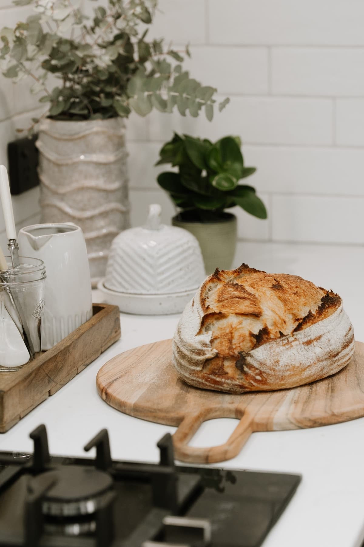 A loaf of sourdough bread sitting on a wooden board on a white counter top. There are some eucalyptus stems in a vase behind the loaf, as well as a dish of butter and another green plant.