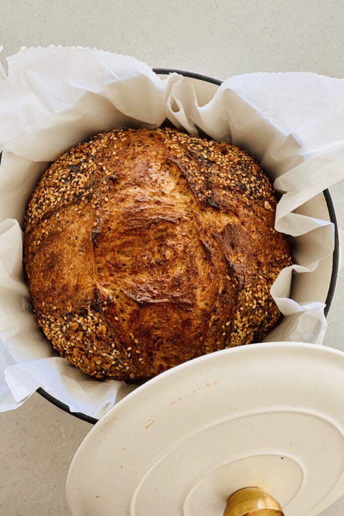 Loaf of multigrain sourdough bread sitting in a cream enamel dutch oven. There is white parchment paper around the baked sourdough loaf.