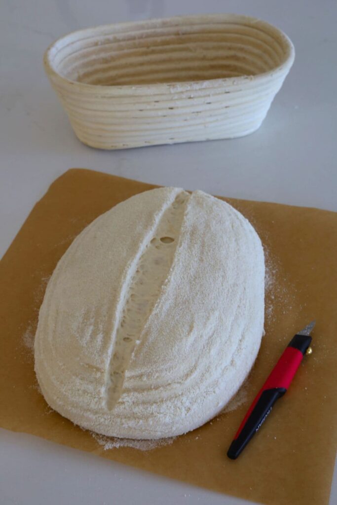 A red handled blade or lame sitting next to a loaf of sourdough that has been scored with a straight line down the centre.