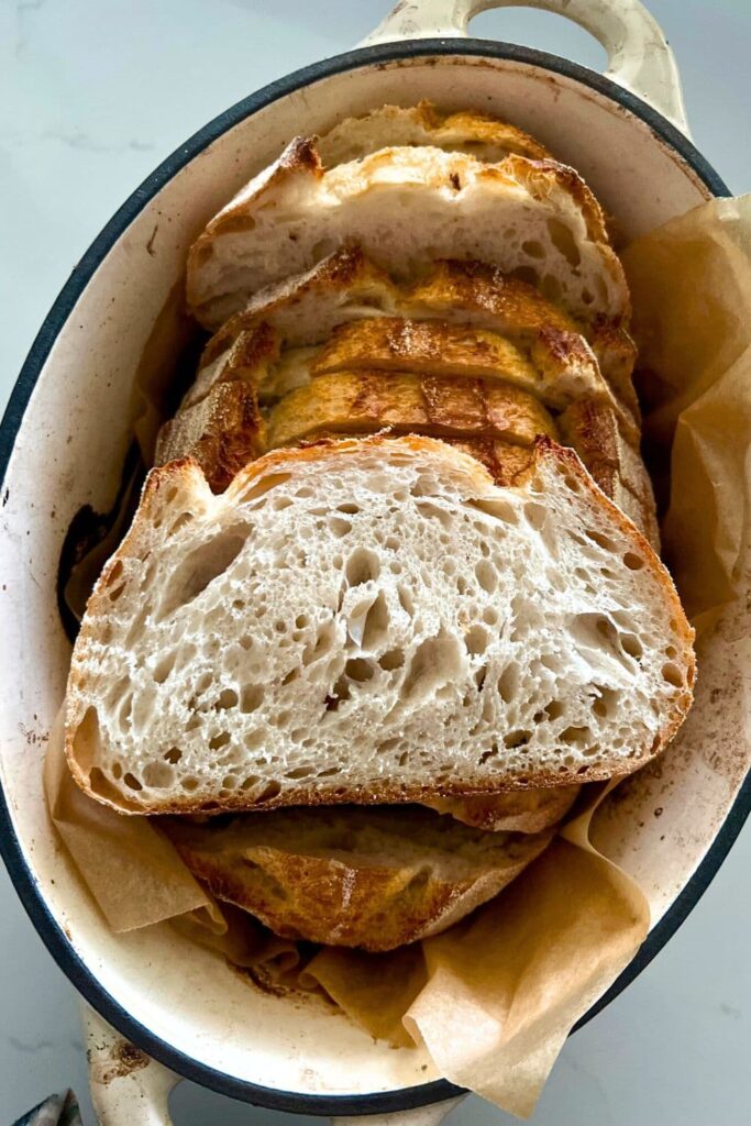 Sourdough Glossary - A loaf of sourdough bread sitting in an oval Dutch Oven. A slice of the sourdough bread has been sat on top so you can see the "crumb shot" or inside of the loaf.