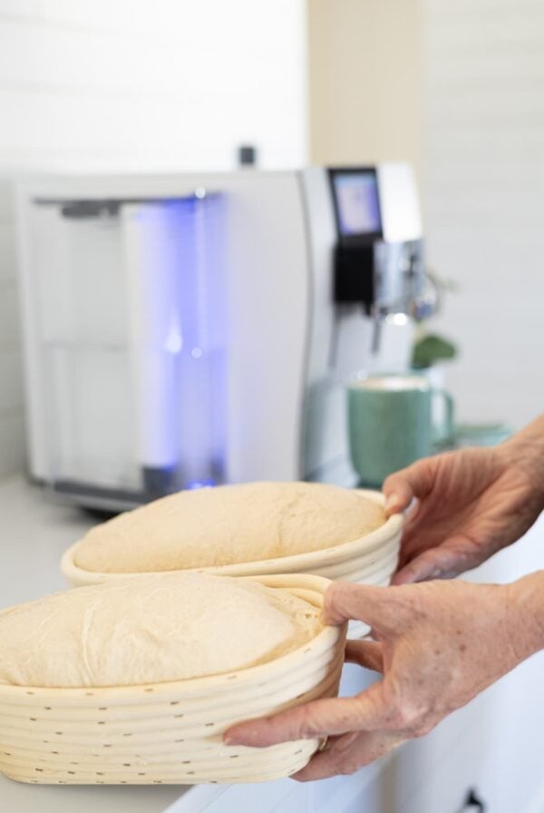 A photo of two bannetons of sourdough being picked up in front of a coffee machine.