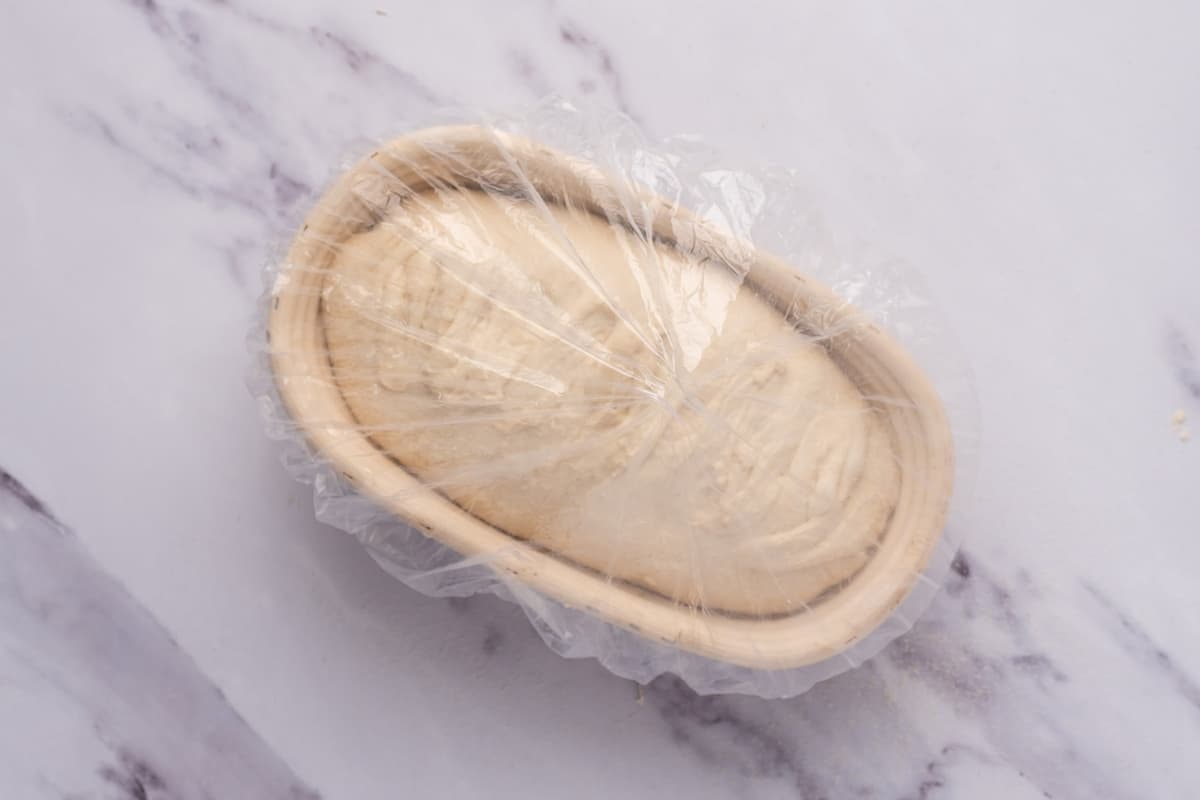 A banneton containing sourdough that has been cold fermented with a plastic cover over the top.