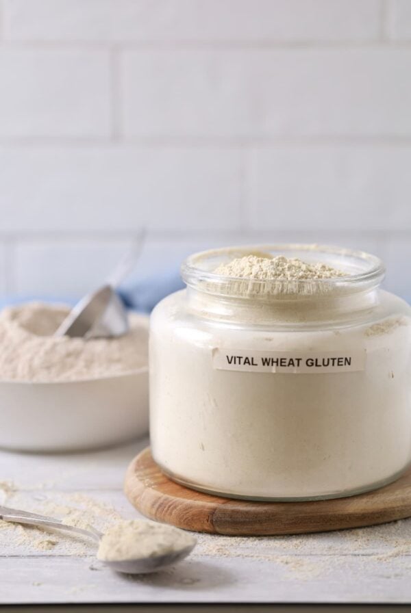A large glass jar filled to the brim with vital wheat gluten. There is a spoon holding some vital wheat gluten sitting in front of the jar.