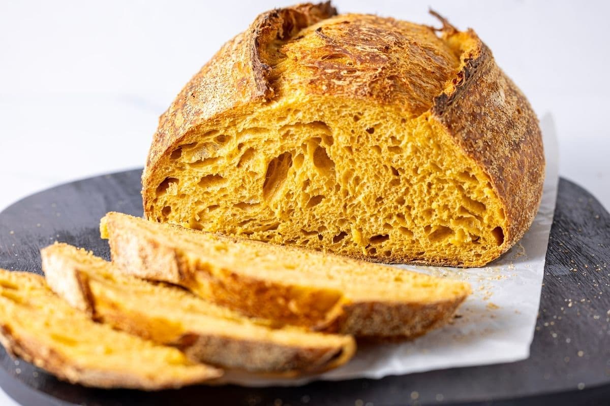 A close up horizontal image of a cut loaf of artisan pumpkin sourdough loaf. You can clearly see the soft, open crumb of the cut face of the loaf. There are some slices laid out in front of the loaf which are slightly out of focus.