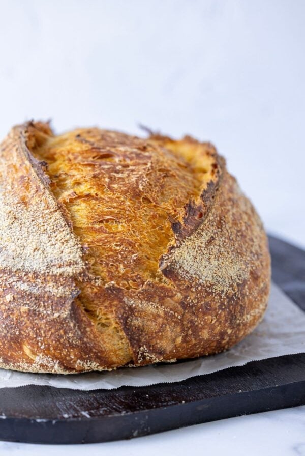 A vertical image showing a very close up image of a whole artisan pumpkin sourdough loaf. The loaf is sitting on a dark wooden board with a piece of white parchment paper sitting under the loaf.