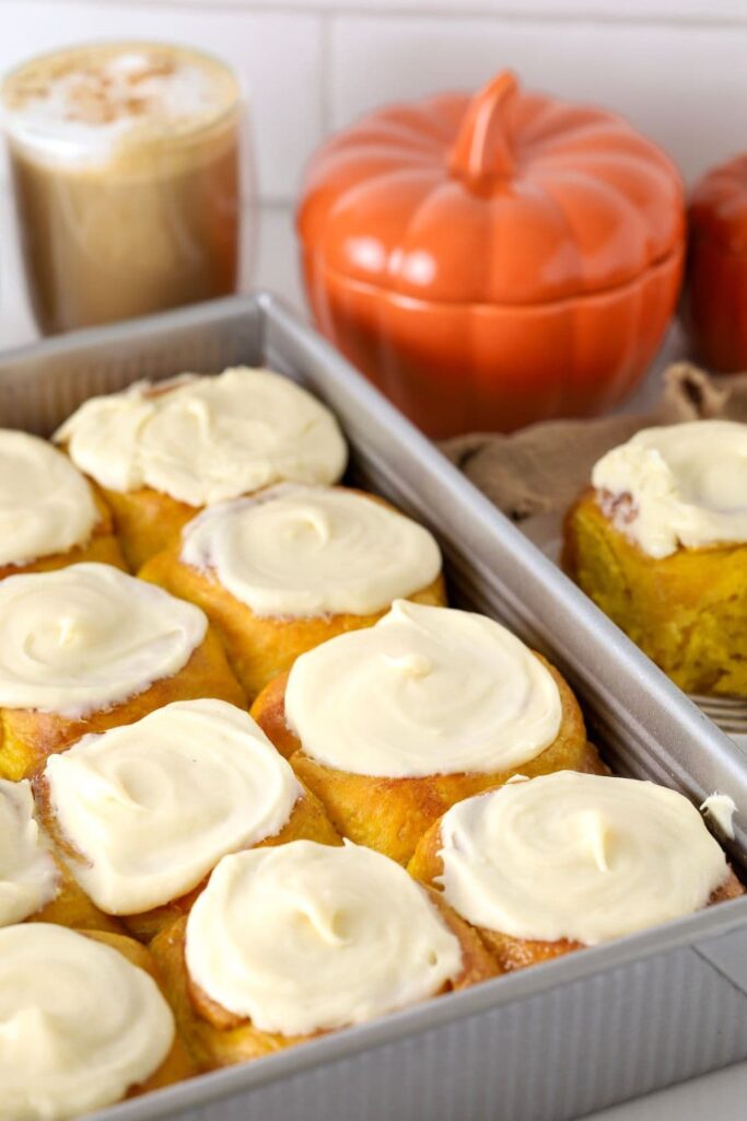 A photo of a tray of sourdough pumpkin cinnamon rolls topped with vanilla cream cheese frosting. You can see a latte in the background an orange pumpkin canister as well.