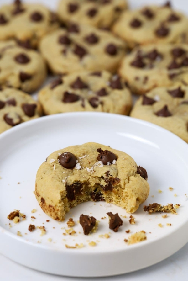 A sourdough chocolate chip cookie sitting on a white plate. There is a bite taken out of the cookie. There are lots of other cookies in the background of the white plate.