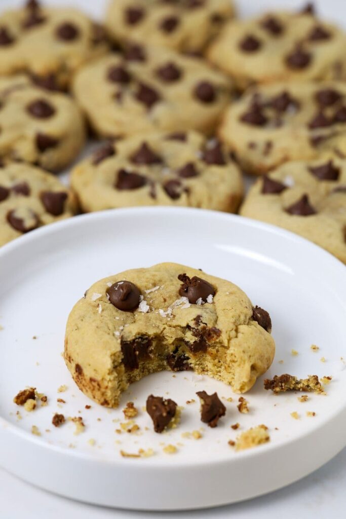 A sourdough chocolate chip cookie sitting on a white plate. There is a bite taken out of the cookie. There are lots of other cookies in the background of the white plate.