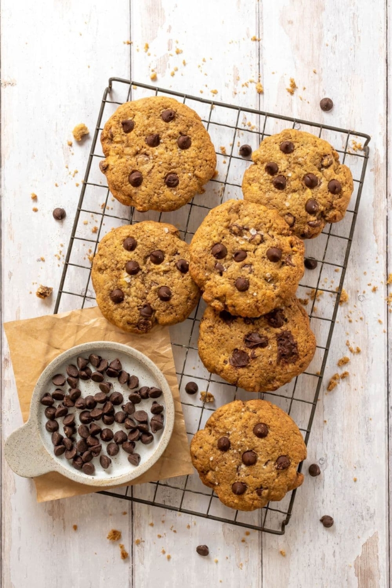 Sourdough chocolate chip cookies piled onto a black wire cooling rack. There is a bowl of chocolate chips sitting in the bottom right corner of the rack.