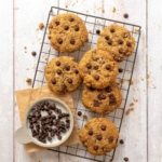 Sourdough chocolate chip cookies piled onto a black wire cooling rack. There is a bowl of chocolate chips sitting in the bottom right corner of the rack.