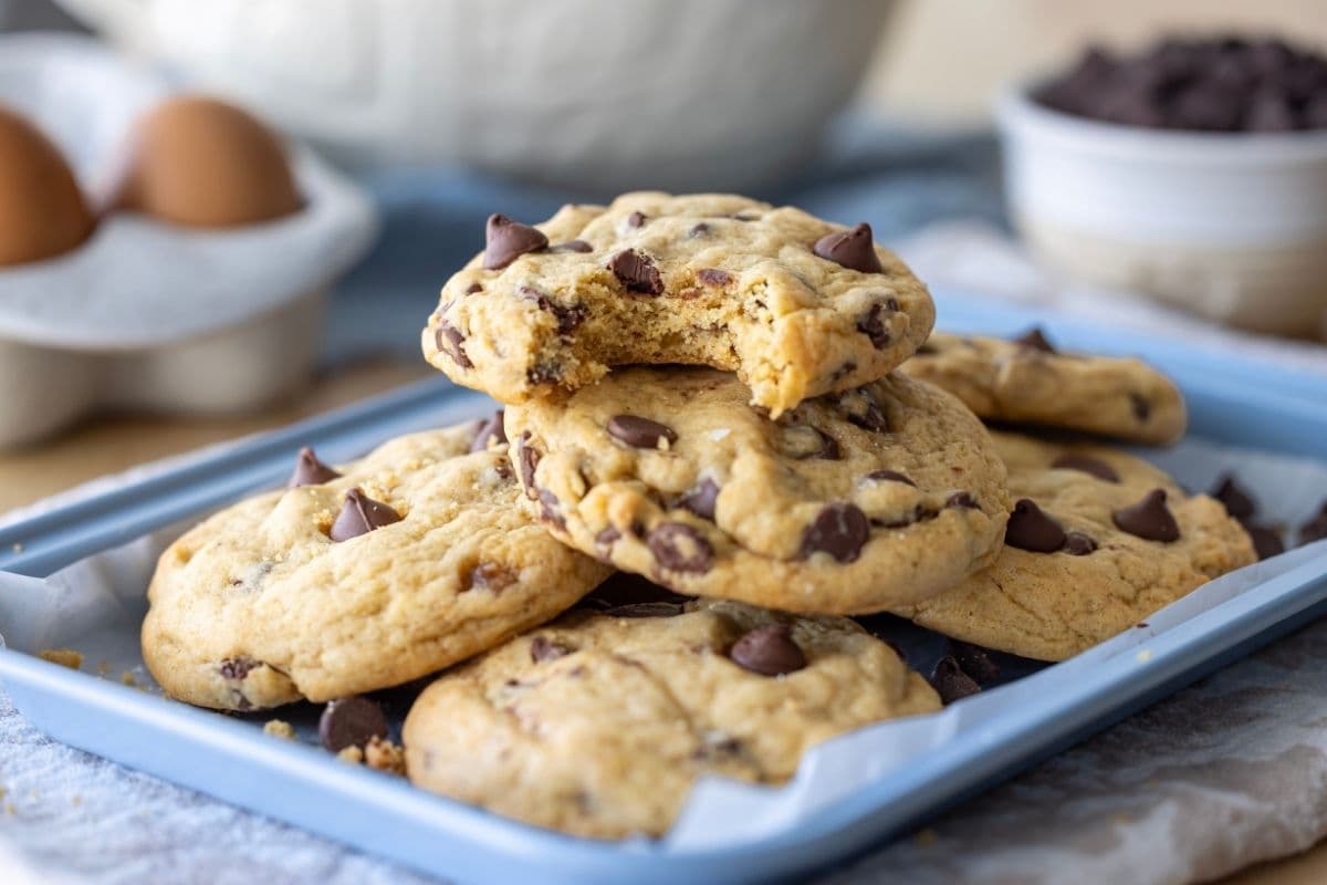A stack of sourdough discard chocolate chip cookies sitting on a pale blue baking tray. The cookie on the top has a bite taken out of it.