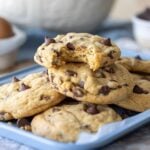 A stack of sourdough discard chocolate chip cookies sitting on a pale blue baking tray. The cookie on the top has a bite taken out of it.