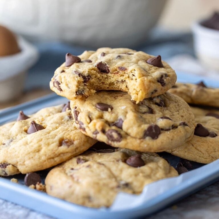 A stack of sourdough discard chocolate chip cookies sitting on a pale blue baking tray. The cookie on the top has a bite taken out of it.