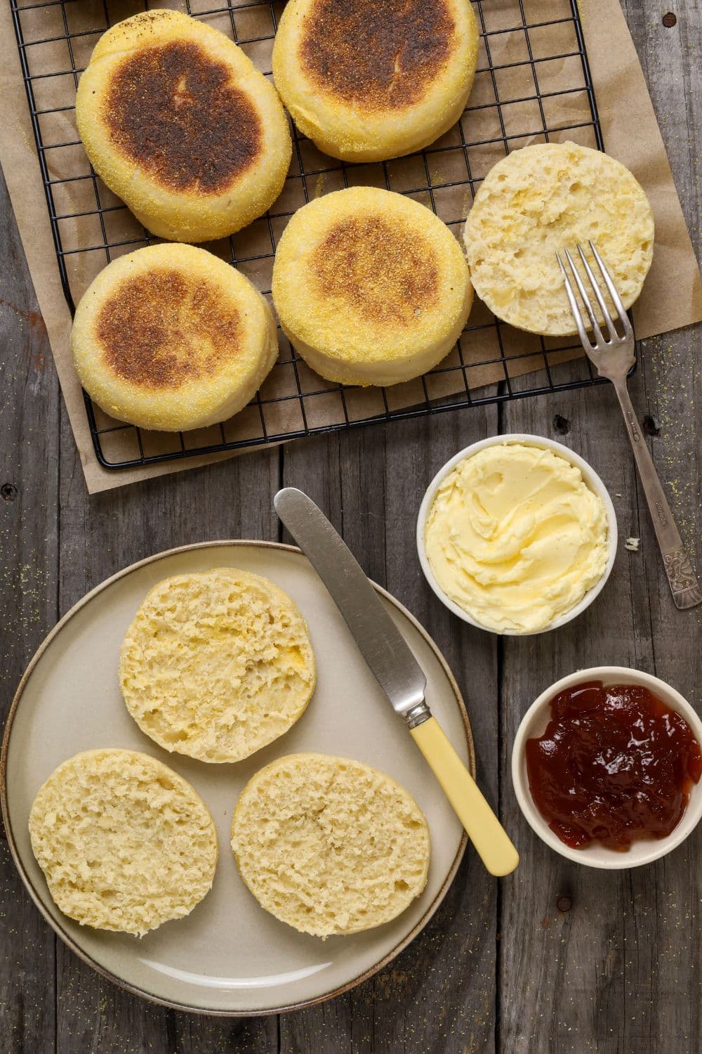 A flat lay photo taken from above showing sourdough English muffins set out for breakfast. Some of the muffins are whole and some have been split open. There are small bowls of butter and jam sitting next to the split English muffins.