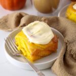 A sourdough pumpkin cinnamon roll sitting on a white plate with a vintage fork. There is a latte colored linen cloth wound around the scene.
