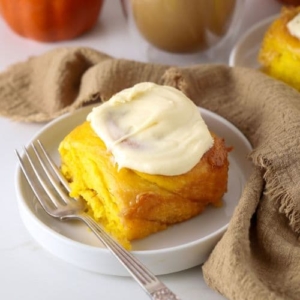 A sourdough pumpkin cinnamon roll sitting on a white plate with a vintage fork. There is a latte colored linen cloth wound around the scene.