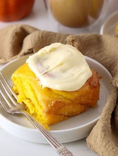 A sourdough pumpkin cinnamon roll sitting on a white plate with a vintage fork. There is a latte colored linen cloth wound around the scene.