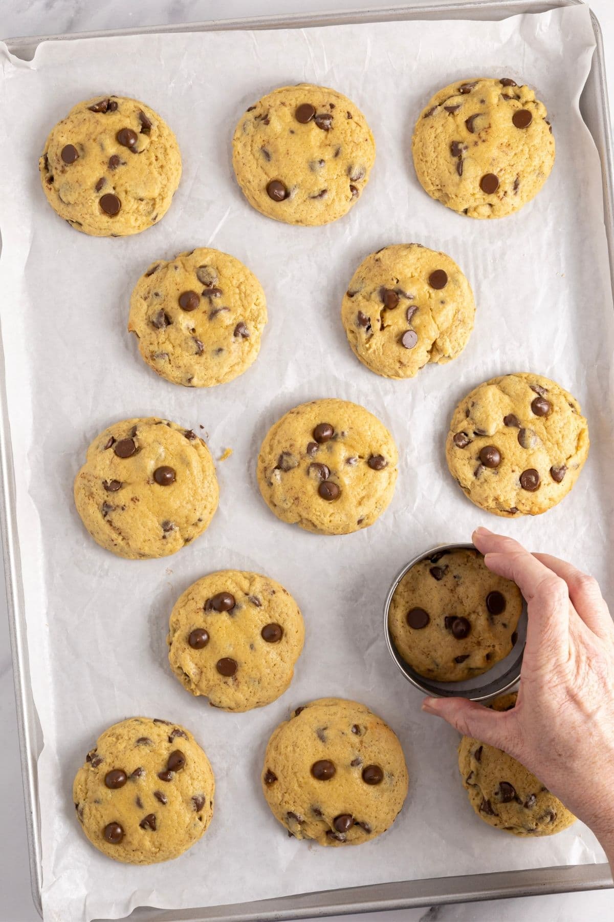 A parchment-lined baking tray filled with just baked sourdough discard chocolate chip cookies. There is a hand swirling the cookies into shape using a round biscuit cutter.