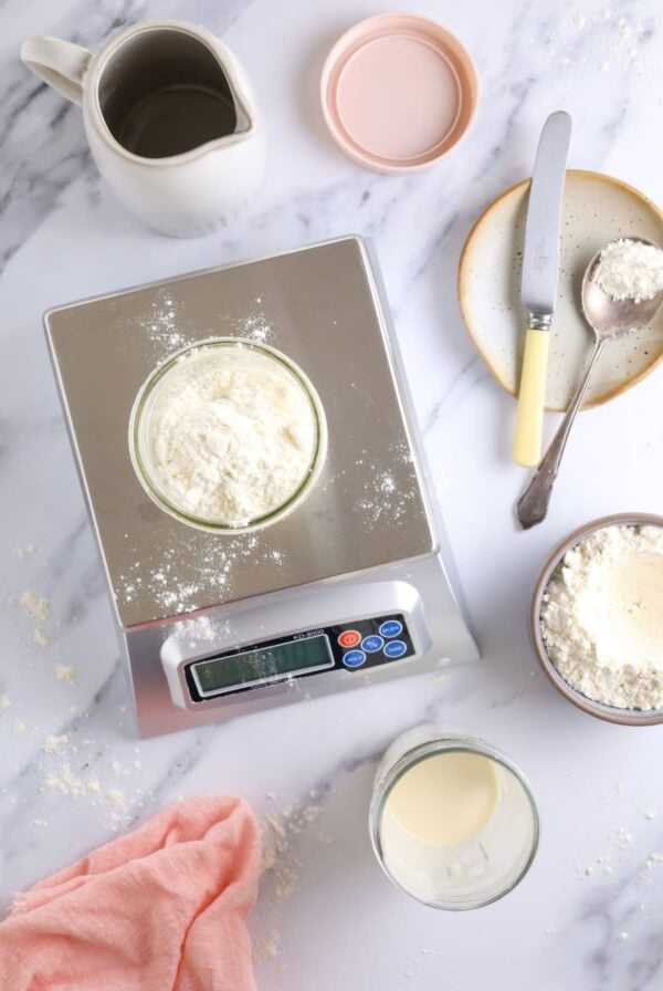 A digital kitchen scale with a jar of sourdough starter sitting on top. Around the digital scale, there is a jug of water, a pink jar lid, a spoon and knife used to stir the starter, as well as a jar of flour and sourdough discard. There is a pale pink cloth in the bottom left corner of the photo.