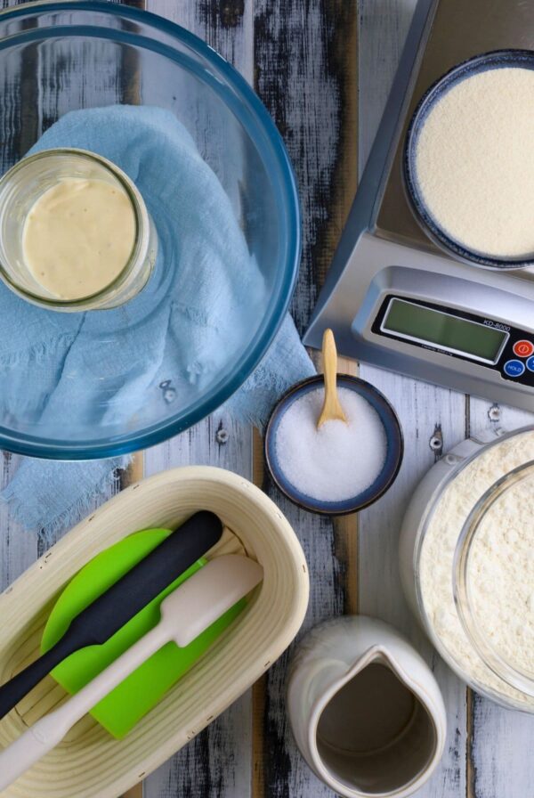 A flat lay of equipment used in sourdough baking including glass bowls, bannetons, digital scale, spatulas etc.