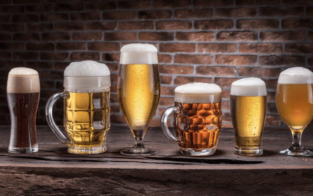 Six glasses of beer sitting in front of a brick wall.