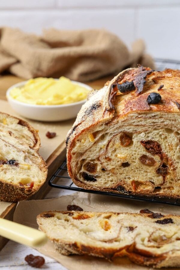 A sourdough fruit loaf full of juicy raisins and dried fruit sitting on a counter top with a dish of butter. The loaf has been cut so you can see the cinnamon swirl and raisins inside.