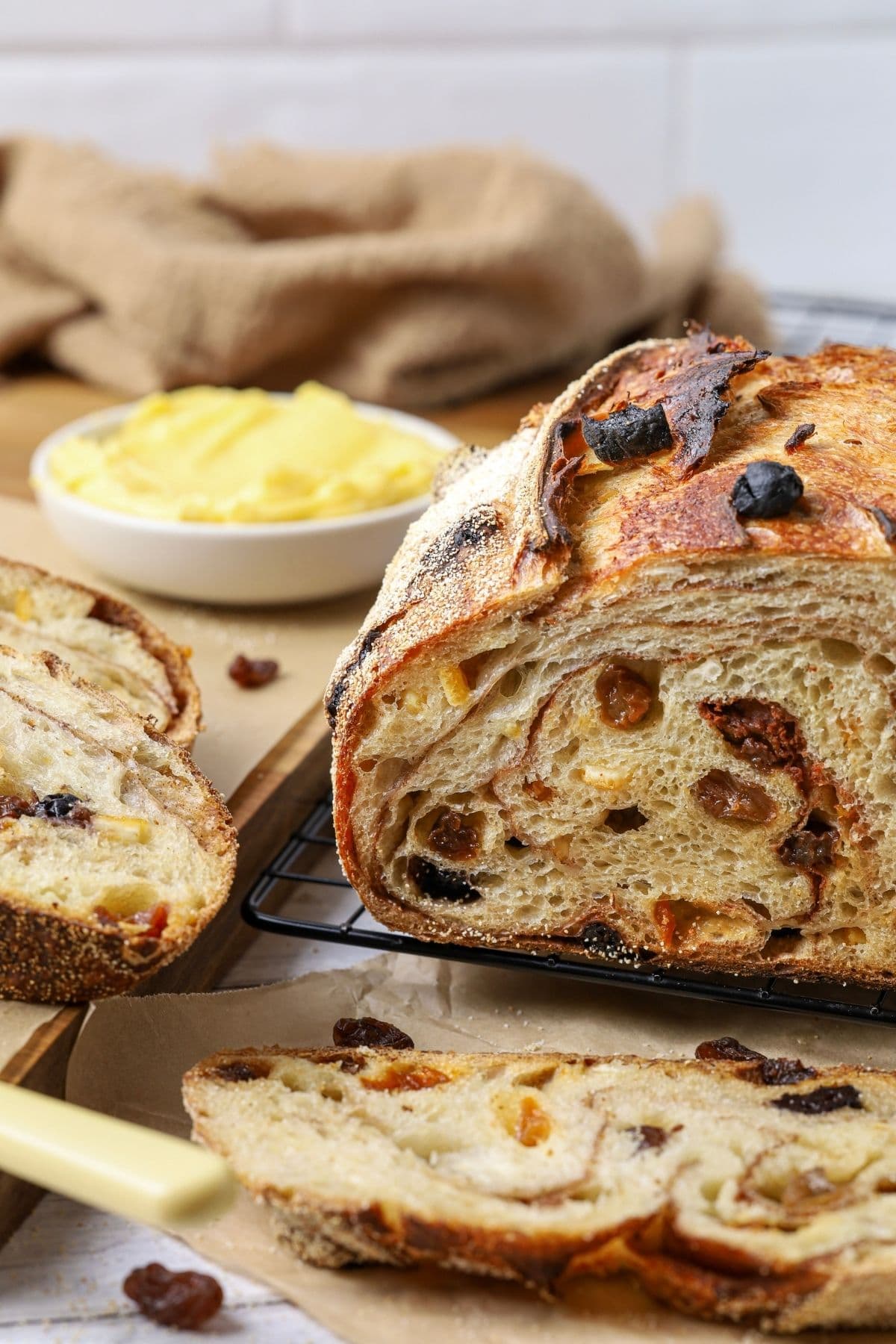 A sourdough fruit loaf full of juicy raisins and dried fruit sitting on a counter top with a dish of butter. The loaf has been cut so you can see the cinnamon swirl and raisins inside.