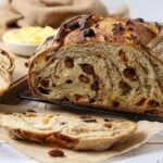 A sourdough fruit loaf full of juicy raisins and dried fruit sitting on a counter top with a dish of butter. The loaf has been cut so you can see the cinnamon swirl and raisins inside.