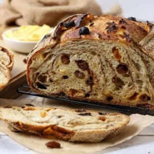 A sourdough fruit loaf full of juicy raisins and dried fruit sitting on a counter top with a dish of butter. The loaf has been cut so you can see the cinnamon swirl and raisins inside.
