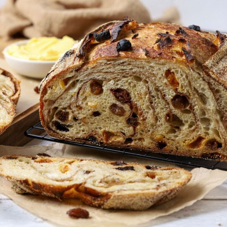 A sourdough fruit loaf full of juicy raisins and dried fruit sitting on a counter top with a dish of butter. The loaf has been cut so you can see the cinnamon swirl and raisins inside.