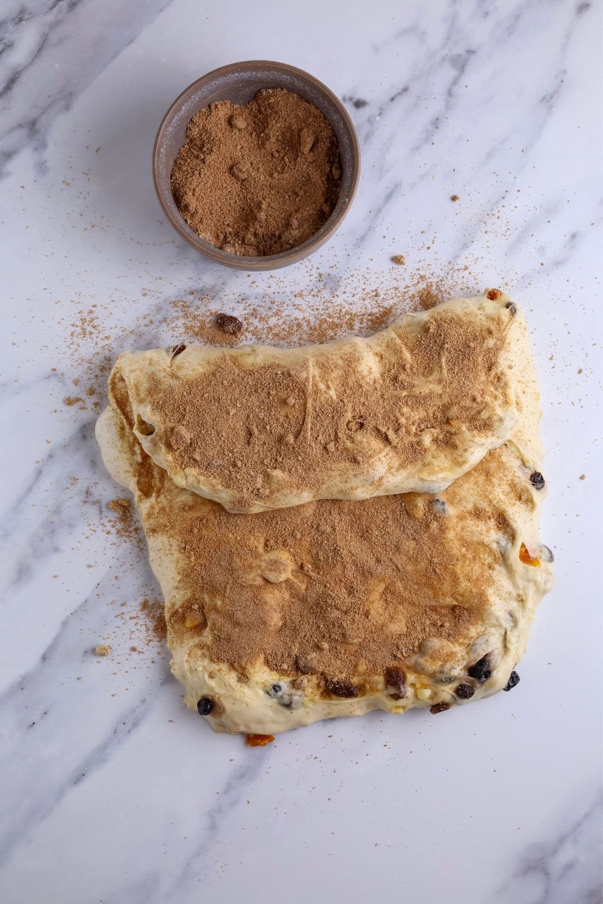 Sourdough fruit loaf dough being shaped. The dough has been pulled out into a rectangle and the cinnamon sugar mixture has been sprinkled on top. The top of the dough has been folded down. 