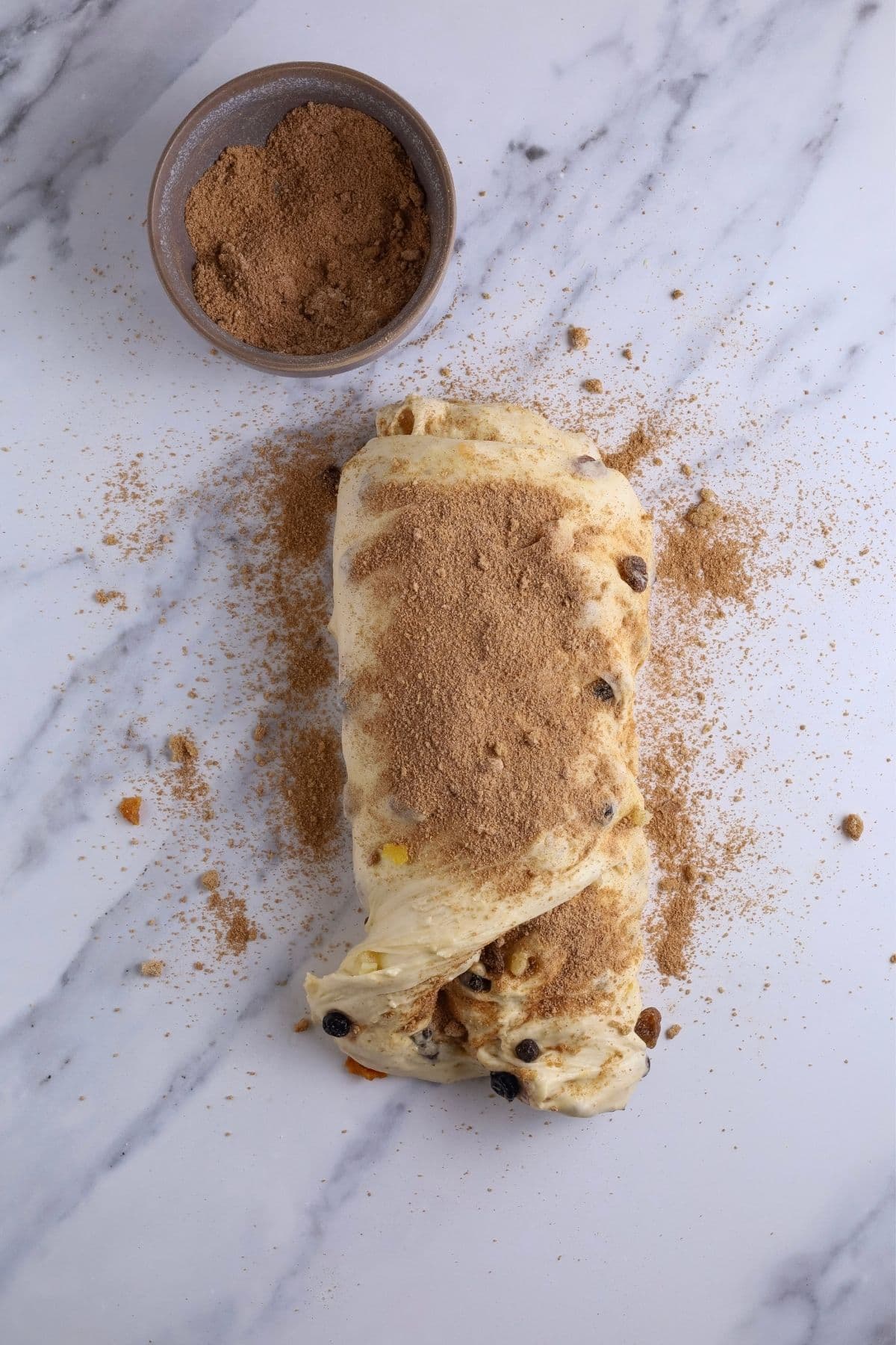 Sourdough fruit loaf dough being shaped. The dough has been pulled out into a rectangle and the cinnamon sugar mixture has been sprinkled on top. The top of the dough has been folded down. And then the other side of the dough has been folded across and more mixture sprinkled on top.