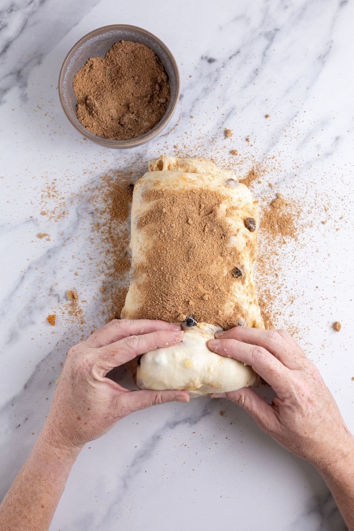Sourdough fruit loaf dough being shaped. The loaf is being rolled up and you can see the hands in the photo doing this. There is cinnamon sugar mixture spilled around the loaf.