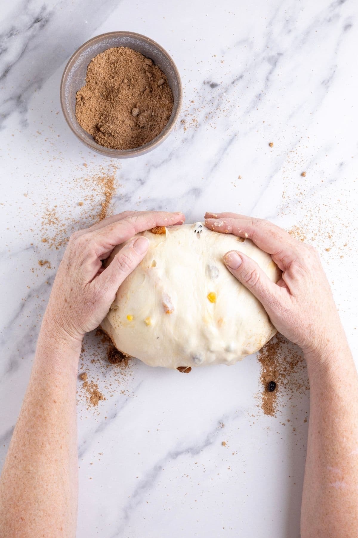 The loaf of sourdough fruit bread shaped into a batard. Hands are pulling the loaf towards them and adding tension to the shaped loaf.
