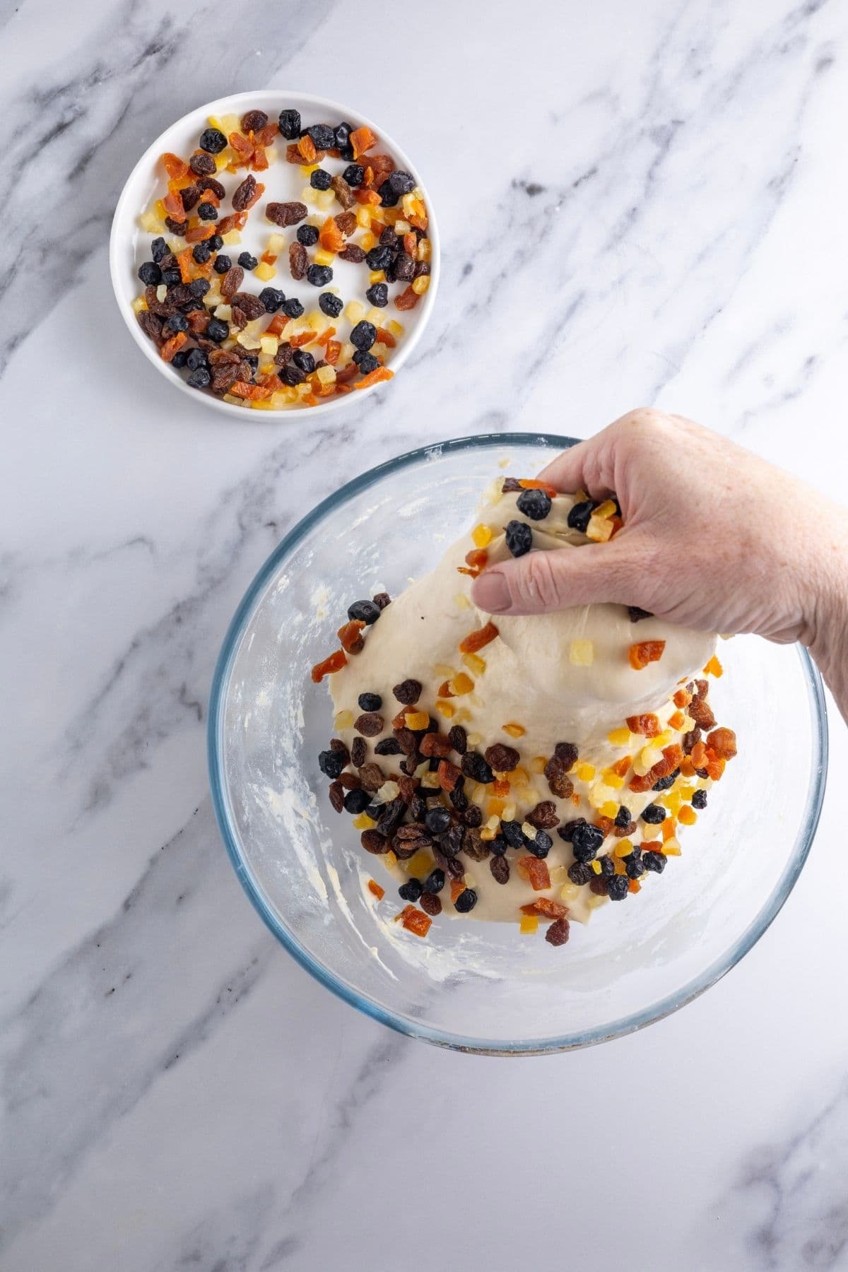A glass bowl of sourdough dough with dried fruit being added to it. Someone is doing a stretch and fold to incorporate the fruit and you can see the hand performing this action in the photo. 
