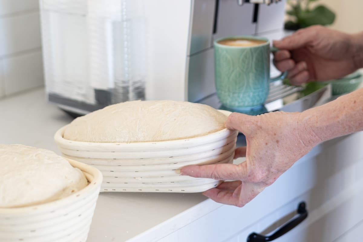 Two bannetons of sourdough sitting next to a coffee machine. There are a pair of hands in the photo and one hand is picking up a banneton and the other hand is picking up a green coffee mug filled with coffee.