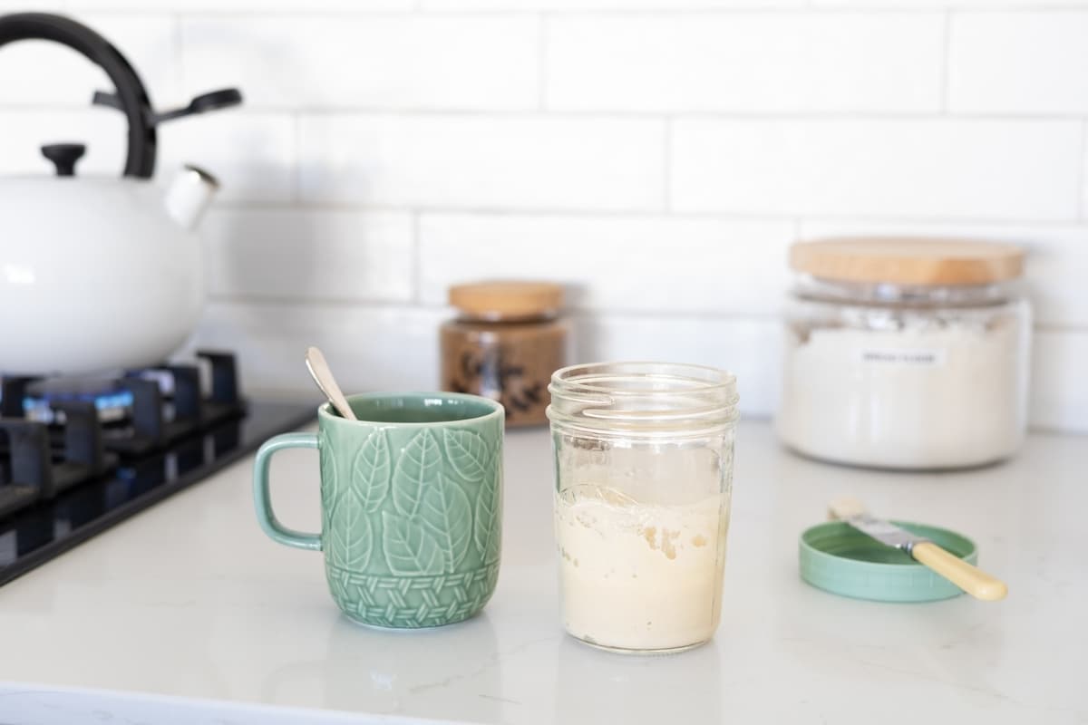 A jar of sourdough starter that has just been fed sitting next to a green coffee mug. There is a kettle being boiled to the left of the coffee mug.