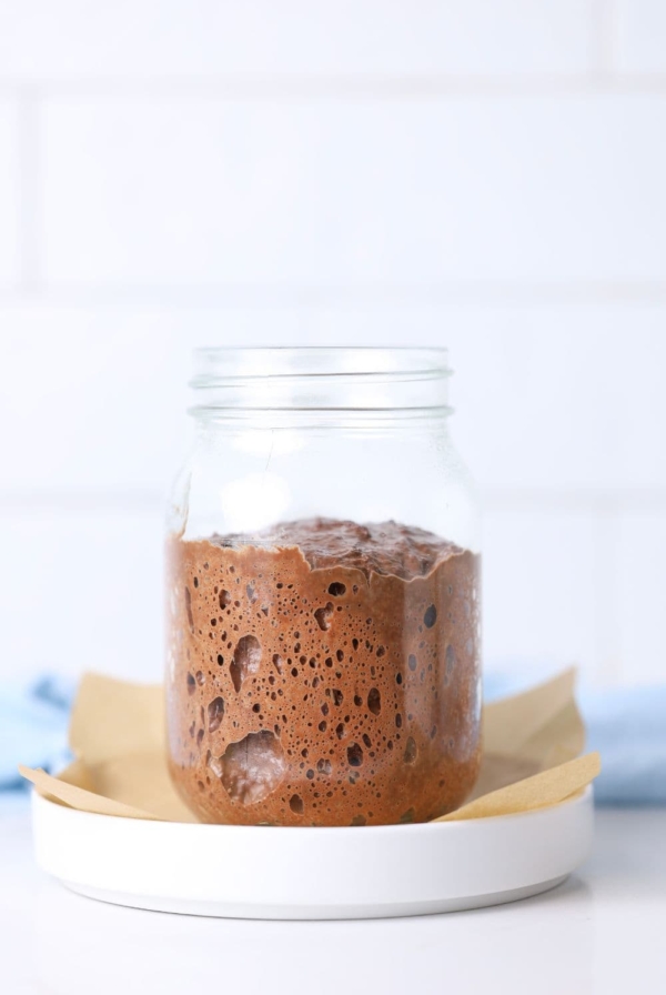 A glass jar containing a chocolate sourdough starter sitting on a white plate with a small piece of parchment paper.