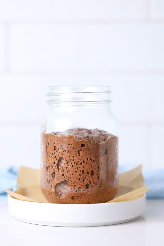 A glass jar containing a chocolate sourdough starter sitting on a white plate with a small piece of parchment paper.