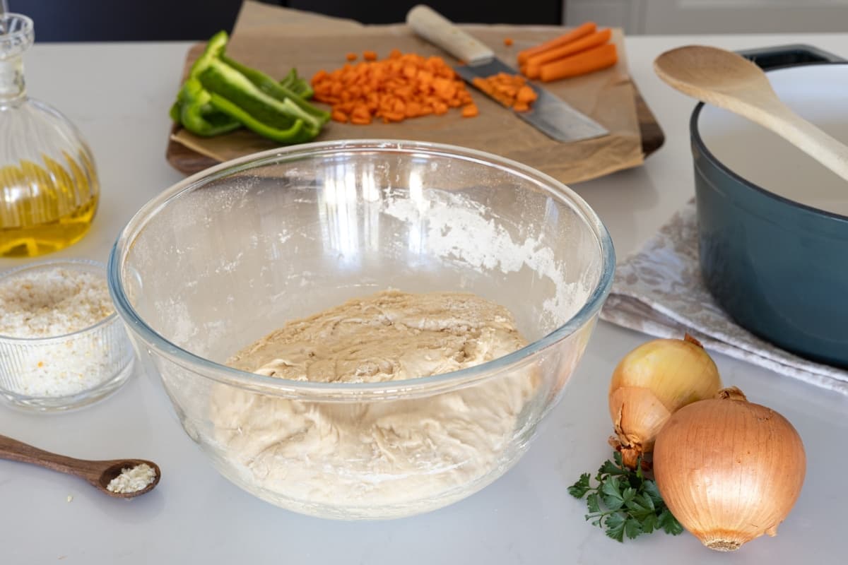 A glass bowl of sourdough sitting in front of a wooden chopping board where orange carrots and green peppers are being chopped up and put into a turquoise colored Dutch oven to the right of the photo.