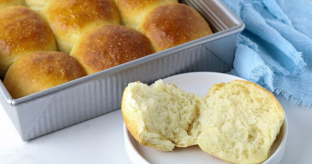 A baking pan of sourdough dinner rolls sitting on a counter top with a dinner roll cut open on a small plate in front of the tray.
