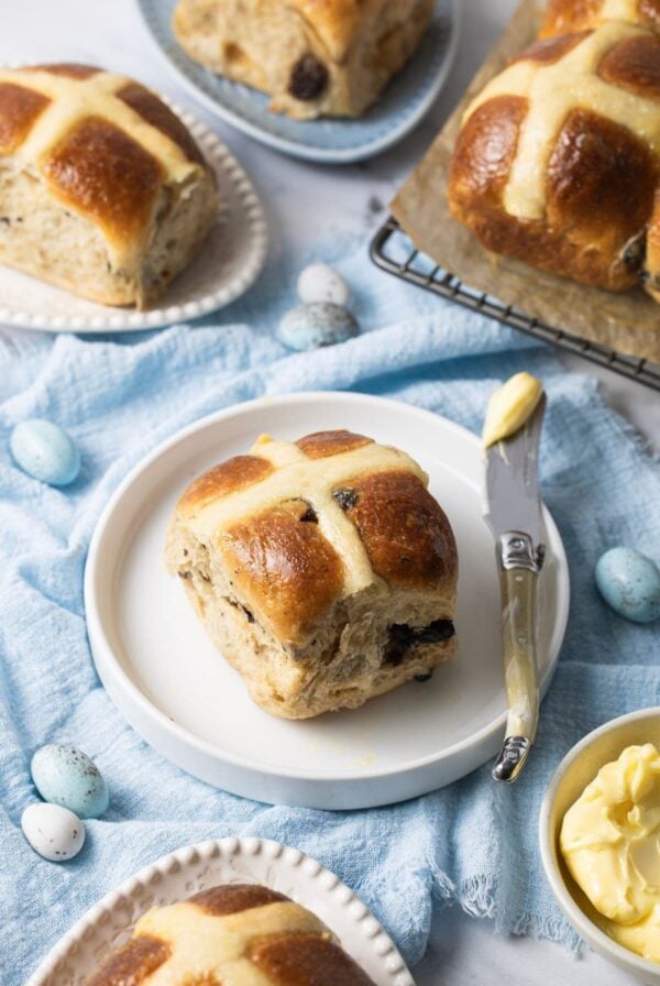 A sourdough hot cross bun sitting on a small white plate surrounded by a pale blue napkin. There are 4 other sourdough hot cross buns in the photos, as well as a small dish of butter.