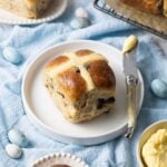 A sourdough hot cross bun sitting on a small white plate surrounded by a pale blue napkin. There are 4 other sourdough hot cross buns in the photos, as well as a small dish of butter.