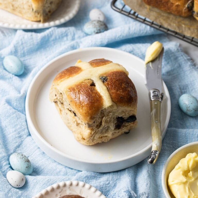 A sourdough hot cross bun sitting on a small white plate surrounded by a pale blue napkin. There are 4 other sourdough hot cross buns in the photos, as well as a small dish of butter.