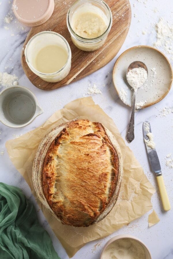 A flat lay showing sourdough mess - there is a large sourdough loaf surrounded by 2 jars of sourdough starter, a messy knife, a spoon with flour on it and the whole scene is dusted with flour mess.