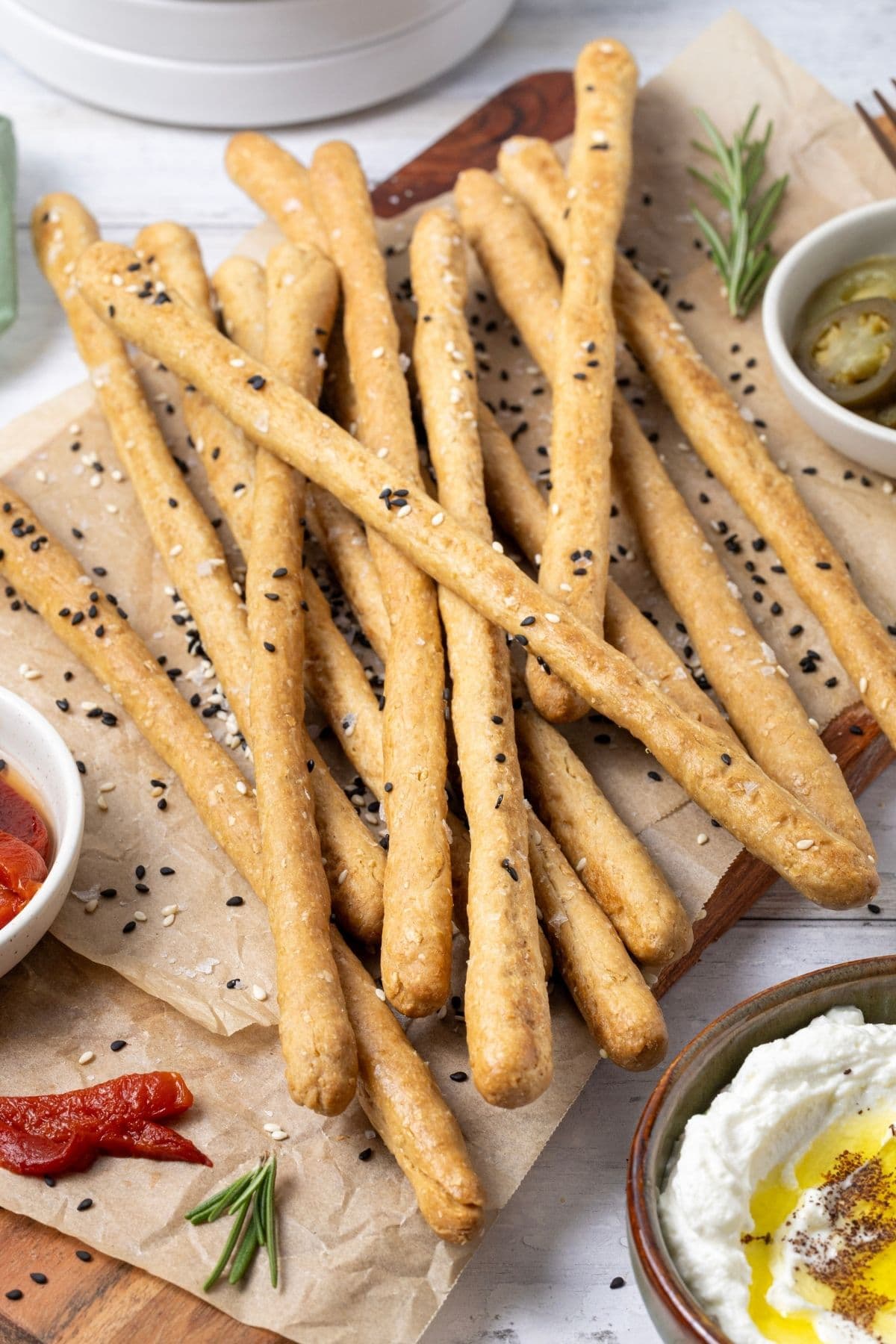A close up photo of sourdough grissini garnished with black and white sesame seeds. The crunchy sourdough breadsticks are piled on a board surrounded by some antipasto type food.