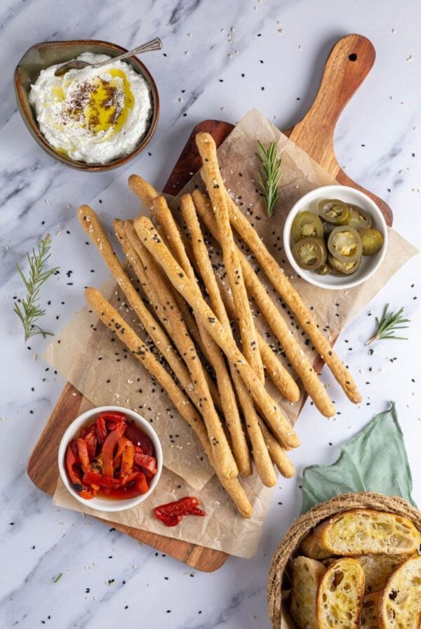 A flat lay image of crunchy sourdough breadsticks displayed on a rectangle serving board. There are some roasted red peppers and jalapenos on the board, as well as some whipped feta dip to the right. The board is garnished with fresh rosemary and there are some sourdough crostini peaking from the right corner of the image.